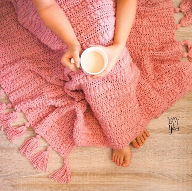 woman under pink crocheted afghan with tassels holding cup of tea